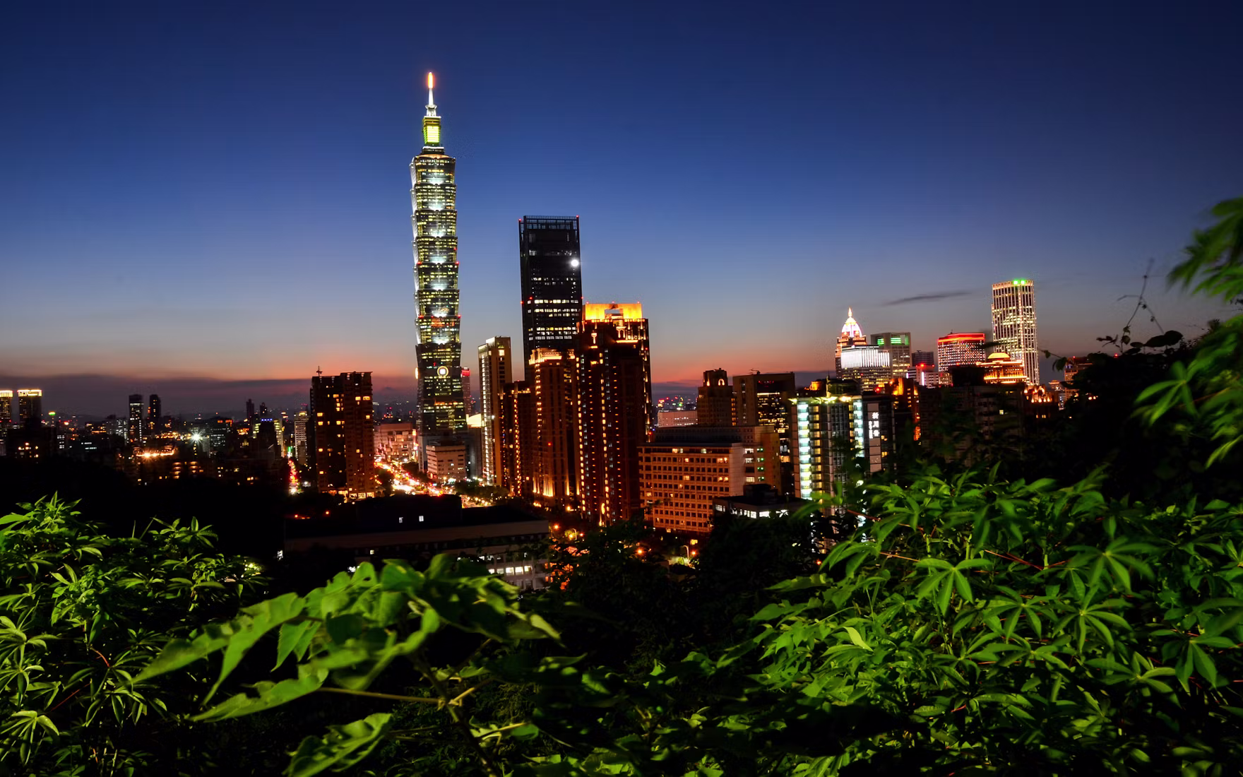 Taipei 101 and the city skyline glowing at golden hour, framed by green hillside leaves